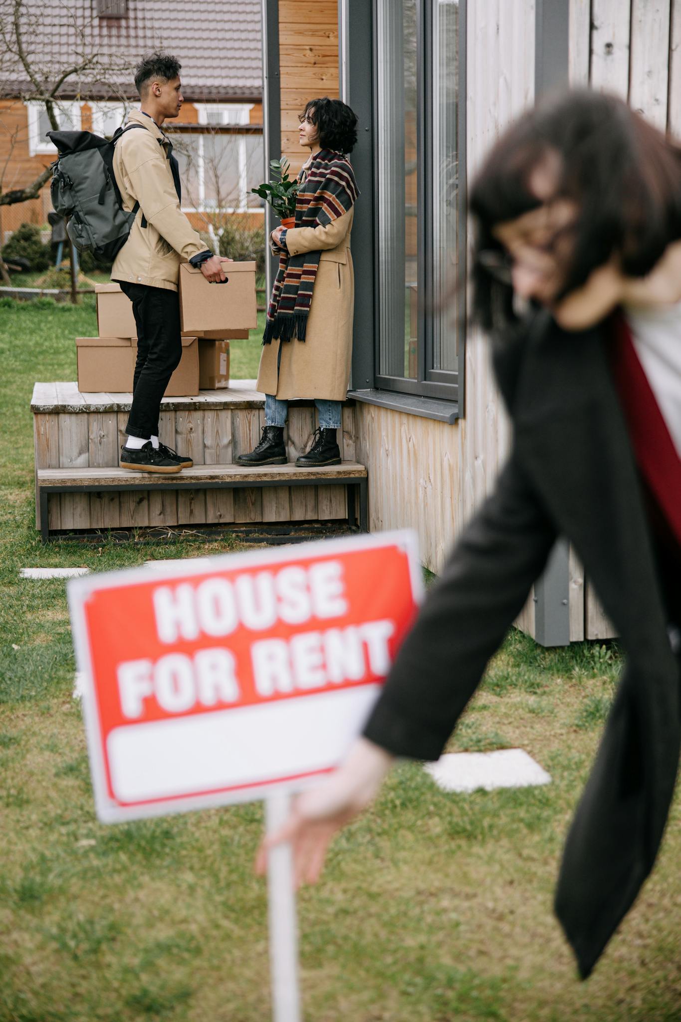 A woman removes a 'House For Rent' sign as new tenants move in, exchanging packages and flowers.